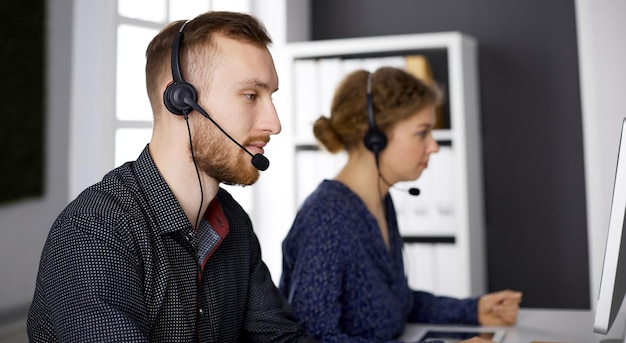 Empresario barbudo hablando por auriculares cerca de su colega mientras está sentado en la oficina moderna. Grupo de personas diversas en el centro de llamadas. Telemarketing y atención al cliente.