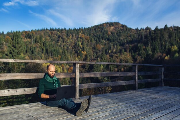 Empresario barbudo sentado en la terraza en las montañas y trabajando en una laptop.