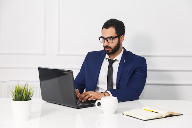 Empresário barbudo usa óculos vestidos de terno azul trabalhar com laptop no interior do escritório branco