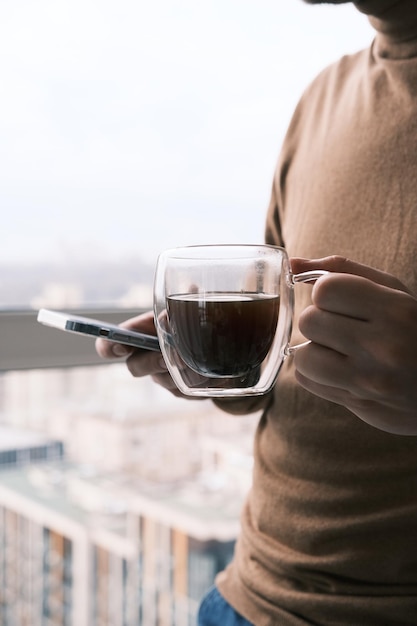 Empresario bebiendo café mirando en un smartphone con una ventana de fondo en una gran ciudad moderna con pensamientos profundos disfrutando de la vista esperando la reunión para empezar a tomar decisiones de negocios