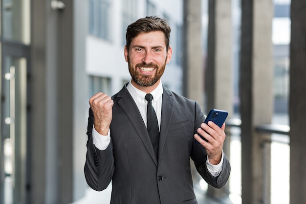 Empresário bem-sucedido e feliz grita de alegria e satisfação por ter vencido, lê boas notícias de um telefone celular, fora do escritório olha para a câmera