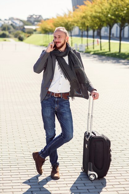 Empresário bem sucedido em traje casual puxa uma mala, corre para o aeroporto e fala em smartphone. Jovem homem caucasiano indo em uma viagem de negócios.