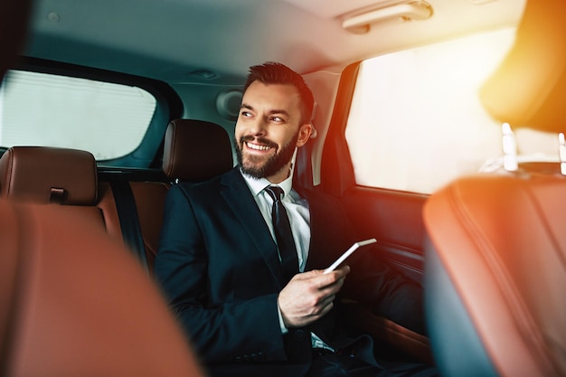 Empresário bonito com telefone na mão sentado no banco de trás do cara do carro olhando para longe