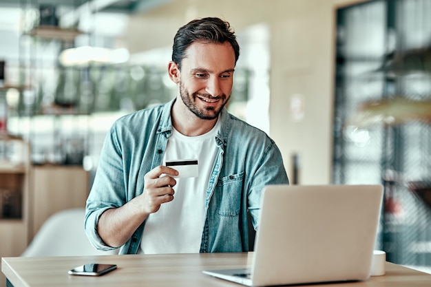 Empresário bonito compras on-line com cartão de crédito Retrato de homem sorridente usando computador portátil no café