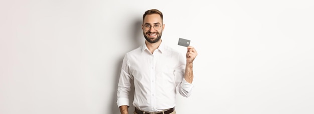 Empresário bonito mostrando seu cartão de crédito parecendo satisfeito em pé sobre fundo branco