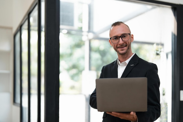 Empresário bonito sorrindo usando laptop em pé no escritório