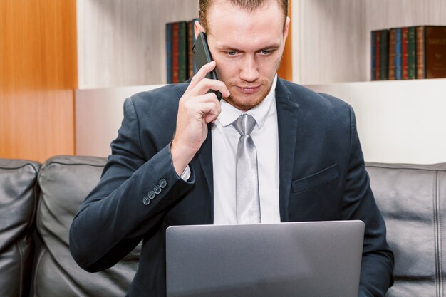 Empresário bonito usando seu smartphone e trabalhando no computador portátil no escritório