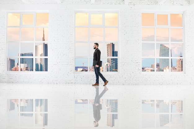 Empresario caminando con un portátil en el interior de la oficina blanca con grandes ventanales con vistas al centro de negocios en la ciudad de Milán