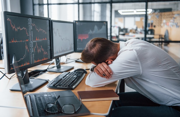 Empresário cansado, dormindo no local de trabalho, inclinando-se sobre a mesa com várias telas. informações de estoque em monitores.