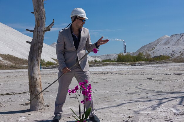 El empresario con un casco protector blanco contra el fondo de la zona industrial protege una flor de orquídea. Concepto ecológico de protección del medio ambiente.