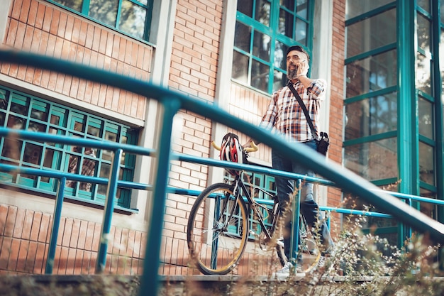 Foto empresário casual vai trabalhar de bicicleta. ele está falando no telefone inteligente e empurrando a bicicleta.