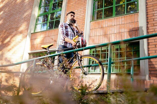 Empresário casual vai trabalhar de bicicleta. Ele está falando no telefone inteligente e empurrando a bicicleta.