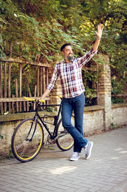 Empresario casual yendo a trabajar en bicicleta. Está caminando al lado de la bicicleta y saludando con la mano.