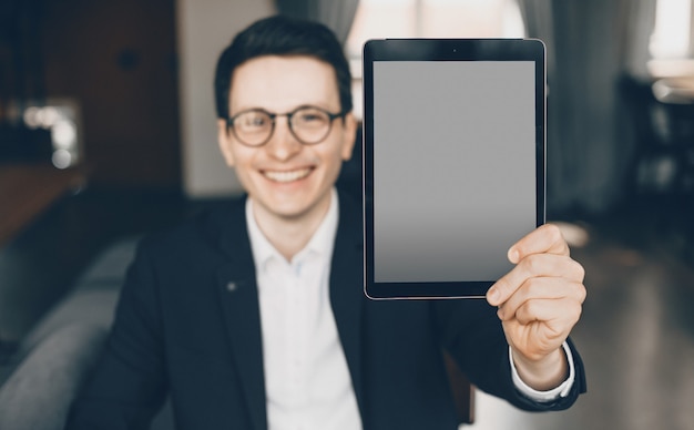 Foto empresário caucasiano sorridente, vestido de terno, mostra um tablet com espaço em branco