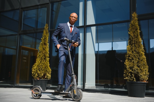 Empresário com scooter elétrico em frente ao edifício empresarial moderno, falando no telefone.