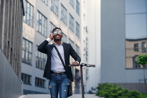 Empresário com scooter elétrico perto de edifício comercial moderno falando ao telefone