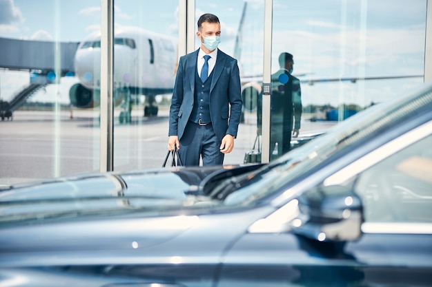 Empresário com uma mala na mão em pé perto da porta de vidro de um aeroporto e olhando para o carro na frente dele. máscara médica no rosto