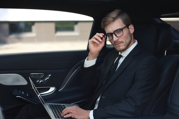 Empresário confiante e pensativo mantendo a mão nos óculos enquanto está sentado no carro de luxo e usando seu laptop.