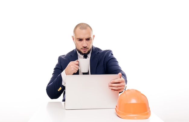 Un empresario constructor cansado con un casco naranja mira la pantalla del portátil y estudia el proyecto de construcción. Se sienta a la mesa y toma café, nervioso