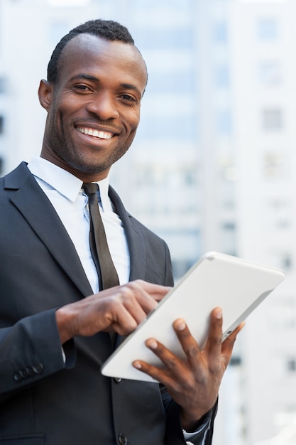 Empresário da era digital. Africano jovem alegre em trajes formais, trabalhando em um tablet digital e sorrindo ao ar livre