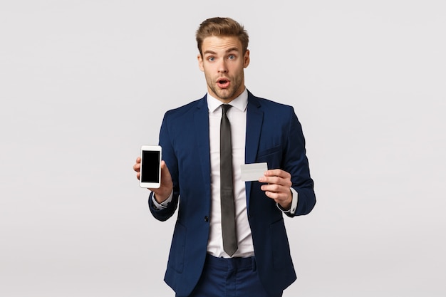 Empresário de bonito chocado e impressionado no elegante terno azul clássico, segurando o cartão de crédito e smartphone, mostrando a tela do celular, diga uau e olhar atônito, promova o app financeiro