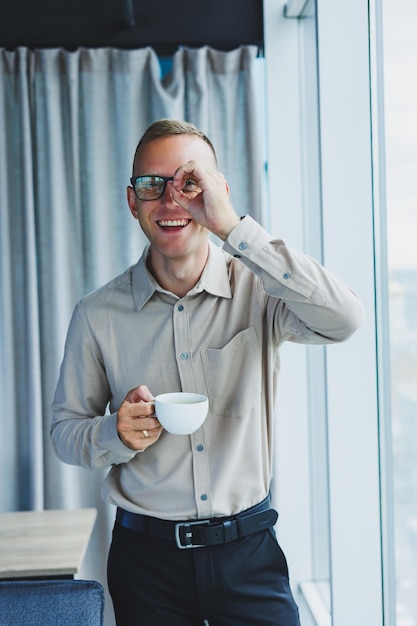 Empresário de escritório moderno ou gerente bebendo café pela janela sorrindo Retrato de programador engenheiro de TI bem sucedido usando óculos trabalhando no escritório