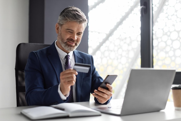 Empresário de meia idade bancário on-line usando smartphone e cartão de crédito no escritório