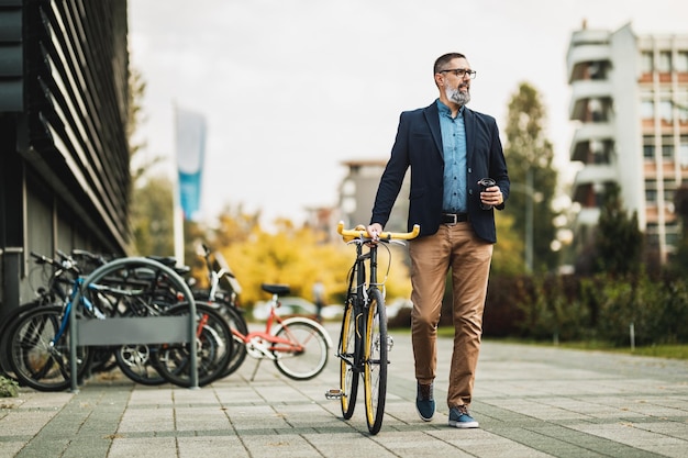 Empresário de meia-idade bem sucedido com uma xícara de café para ir e com bicicleta ao lado dele, caminha em frente ao distrito de escritórios.