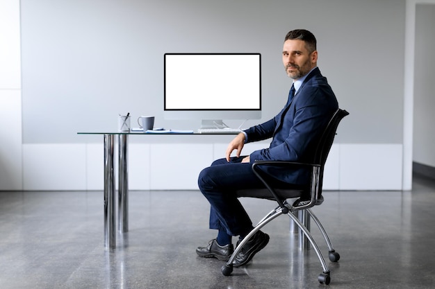 Empresário de meia-idade confiante em terno sentado na mesa com monitor de computador com tela em branco na maquete do escritório