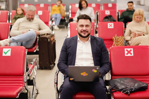 Empresário de meia-idade sorridente em trajes formais usando laptop enquanto está sentado no saguão de um aeroporto moderno contra outras pessoas esperando o voo