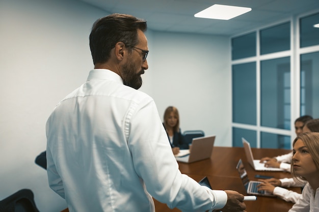 Empresário de pé, de costas para o primeiro plano, discutindo com a equipe que trabalha na mesa grande