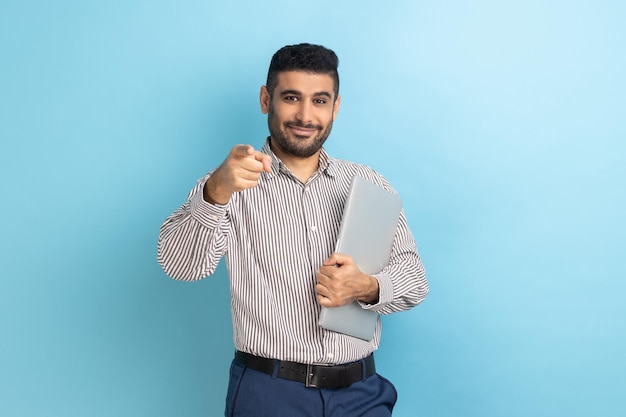 Empresário de pé segurando laptop ou pasta fechada tem expressão positiva apontando para a câmera