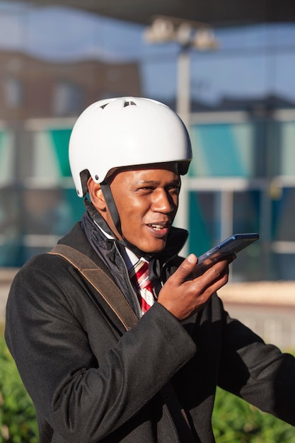 Empresário de pele escura em um capacete de scooter segura o telefone e sorri enquanto fala em mensagens de voz