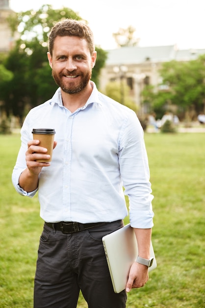 empresário de sucesso com camisa branca, em pé com café para viagem e laptop prateado durante um passeio no parque