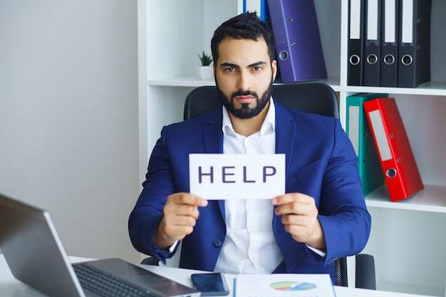 Empresário de terno e gravata, sentado na mesa do escritório, trabalhando no laptop computador pedindo ajuda segurando placa de papelão