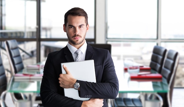 Foto empresário de terno posando com seu laptop contra a sala de reuniões corporativa vazia