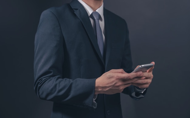 Empresário de terno preto e gravata com segurando o smartphone em fundo preto.