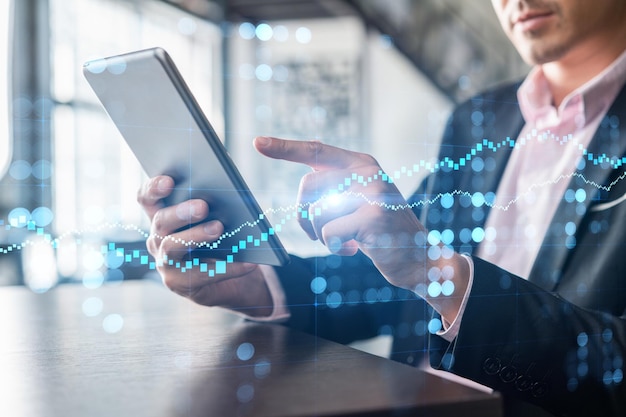 Empresário de terno usando dispositivo tablet para otimizar a estratégia de negociação no fundo de finanças corporativas holograma gráfico Forex sobre fundo de escritório panorâmico moderno