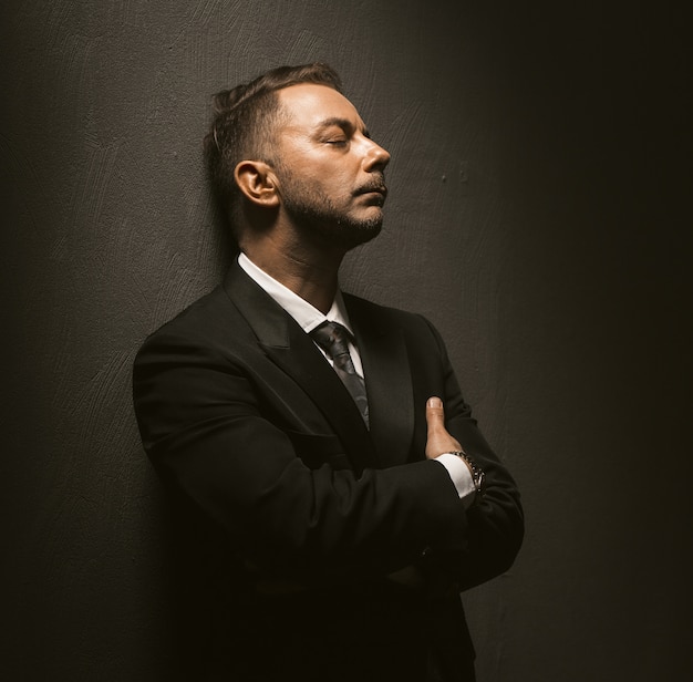 Empresario deprimido se encuentra en cuarto oscuro. El hombre triste se para con los ojos cerrados y los brazos cruzados, apoyando la espalda contra la pared sobre fondo gris. Imagen tonificada