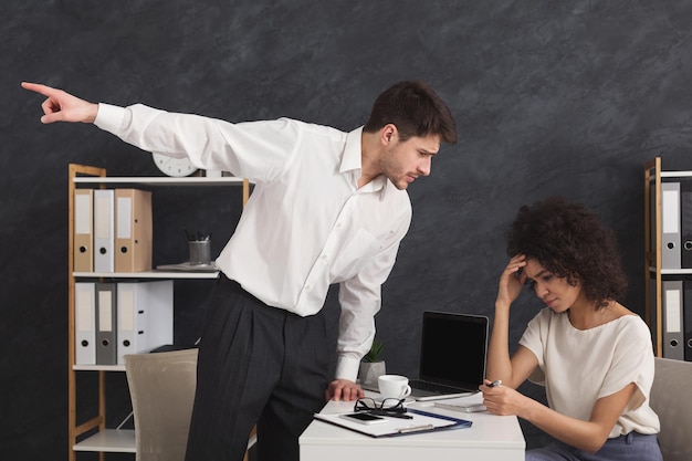 Empresario despidiendo a su secretaria llorando. Mujer afroamericana molesta sentada en el lugar de trabajo, su jefe enojado apunta a la puerta, espacio de copia