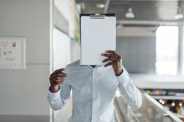 El empresario de ébano se cubre la cara con un cuaderno en el centro comercial. Persona de negocios exitosa, hombre negro en ropa formal, centro comercial