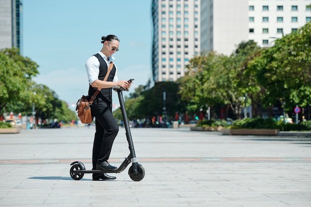 Empresário elegante sorridente com scooter elétrico ao ar livre lendo mensagens de texto no smartphone