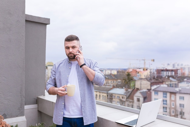 Empresário em roupas casuais relaxando no terraço com vista para a cidade usa o telefone e usa um laptop enquanto bebe café