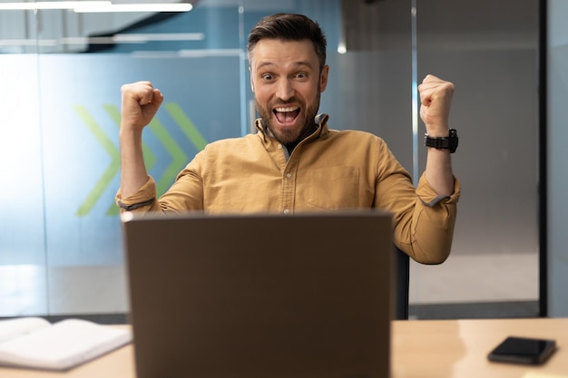 Foto empresario emocional en la computadora portátil gesticulando sí sacudiendo los puños en la oficina