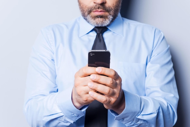 Empresario escribiendo mensajes. Primer plano de hombre maduro confiado en camisa y corbata escribiendo un mensaje en el teléfono móvil mientras está de pie contra el fondo gris