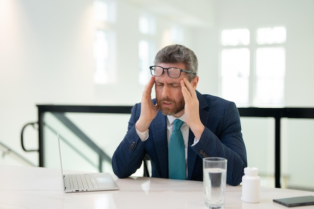 Empresário estressado com dor de cabeça no escritório Empresário cansado está trabalhando horas extras e tem dor de cabeça Homem sobrecarregado de trabalho com laptop no local de trabalho sofre de dor de cabeça enxaqueca por excesso de trabalho