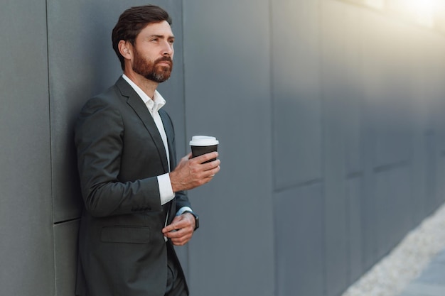 Empresario europeo enfocado en traje bebiendo café cerca del edificio de oficinas durante el descanso