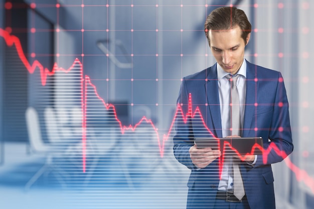 Empresário europeu pensativo atraente usando tablet com grade de gráfico de negócios vermelho caindo no fundo interior do escritório embaçado Recessão de crise e conceito de mercado de ações Dupla exposição