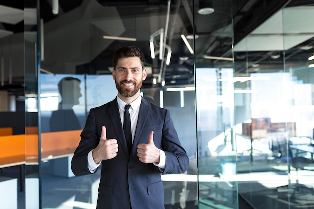 Empresário feliz e bem sucedido olhando para a câmera e sorrindo com aprovação mostrando os polegares no escritório moderno