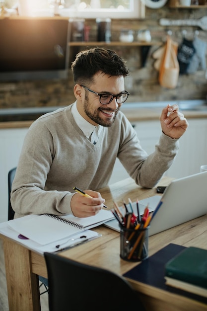 Empresario feliz usando laptop y tomando notas mientras trabaja en casa